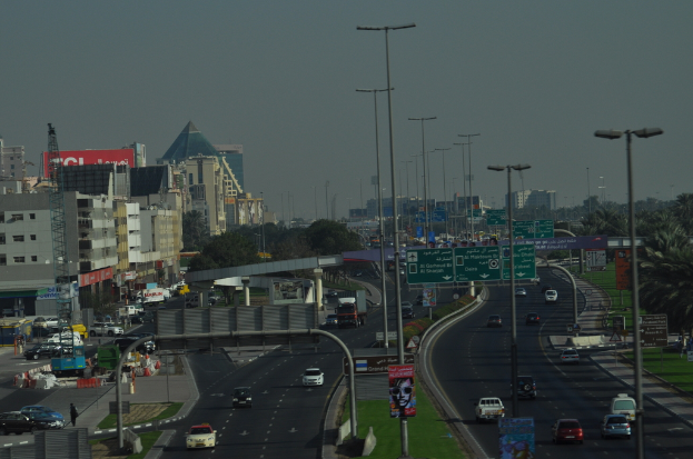 Eine Luftansicht einer Stadt mit Gebäuden, Türmen, Kraftfahrzeugen, Werbetafeln, Schildern, Straßen, Bäumen, Straßenlaternen, Menschen, Verkehrskegeln und Informationsschildern unter einem klaren Himmel.