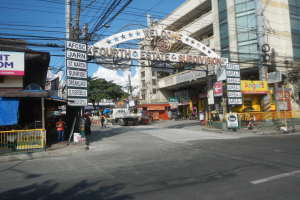 Eine belebte Straße in der Stadt mit fahrenden Fahrzeugen, Füßgängern auf dem Gehweg, einem Tor mit Text, Gebäuden mit Fenstern, Strommästen mit Überlandleitungen, Bäumen und einem bewölkten Himmel, mittig von einem Straßenschild mit der Aufschrift "Fourth Estate Subdivision".