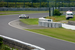 Ein Rennwagen fährt auf einer Rennstrecke mit einem Polizeiwagen im Hintergrund, umgeben von Gras, Pflanzen, Bäumen und einer Umzäunung, mit einem Fahrzeug auf der rechten Seite und Text unten.