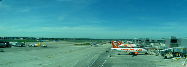 Eine Luftaufnahme eines Flughafens mit Flugzeugen, Gebäuden, Masten, Fahrzeugen, Bäumen und einem Zaun im Vordergrund.
