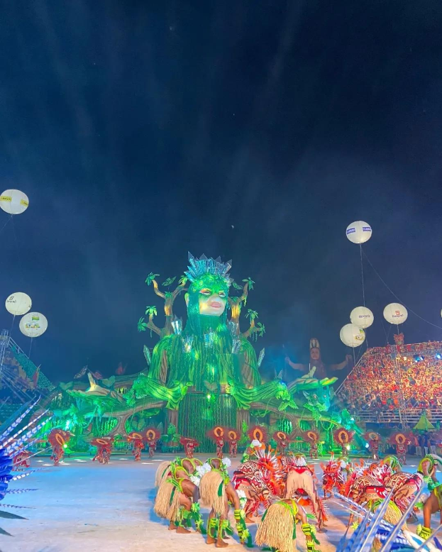 Ein farbenfroher Karneval in Rio de Janeiro, Brasilien, mit Performern in bunten Kostümen auf der Bühne, einer großen Menge im Publikum und einem Himmel voller bunter Lichter und Ballons.