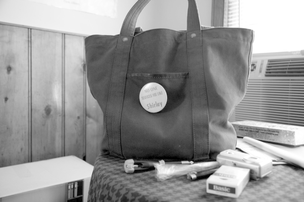 Eine Handtasche mit einer Kamera und einem Zigarettenetui darauf steht auf einem Tisch in einem Raum, mit einer Klimaanlage und einem Fenster darüber; die Tasche hat ein Etikett, auf dem steht: "Niemand bedient Sie wie Shirley."