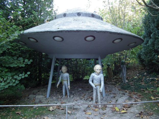 Eine Gruppe von Menschen steht vor einem UFO, umgeben von Bäumen und einem klaren blauen Himmel, mit zerrissenen Blättern und Steinen auf dem Boden und einem Geländer im Vordergrund.