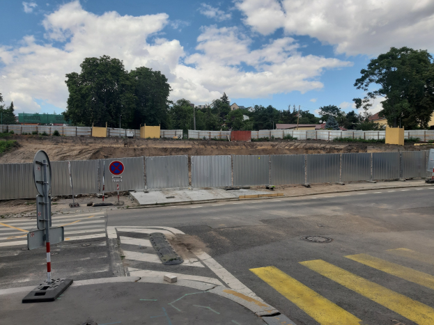 Eine Baustelle mit einer teilweise gebauten Straße, die mit gelben und weißen Streifen markiert ist, ein Metallzaun, ein Schild, Pfosten, Bäume, Gebäude und ein bewölkter Himmel.
