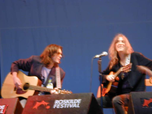 Zwei Männer auf der Bühne mit Musikinstrumenten und einem Mikrofon, mit "ROCK SLIDE FESTIVAL" auf Kisten auf dem Boden.