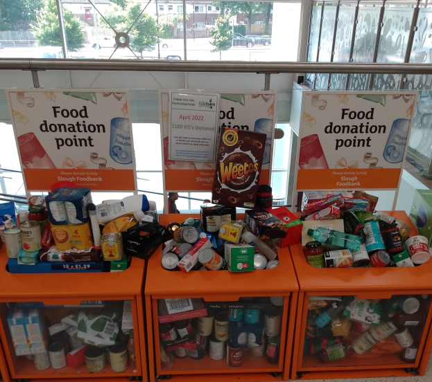 Orange Schränke mit Lebensmitteln wie Flaschen, Dosen und Kartons an einer Spendenstelle, mit Informationsschildern und einer Aussicht auf Fahrzeuge, Bäume, Gebäude und Himmel durch Glasfenster.