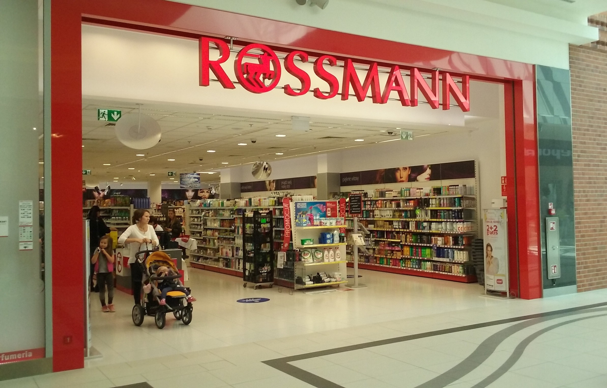 Ein Rossmann-Laden in einem Einkaufszentrum mit Deckenbeleuchtung, Schildern, Menschen und Regalen voller verschiedener Gegenstände, eine Person mit einem Kinderwagen.
