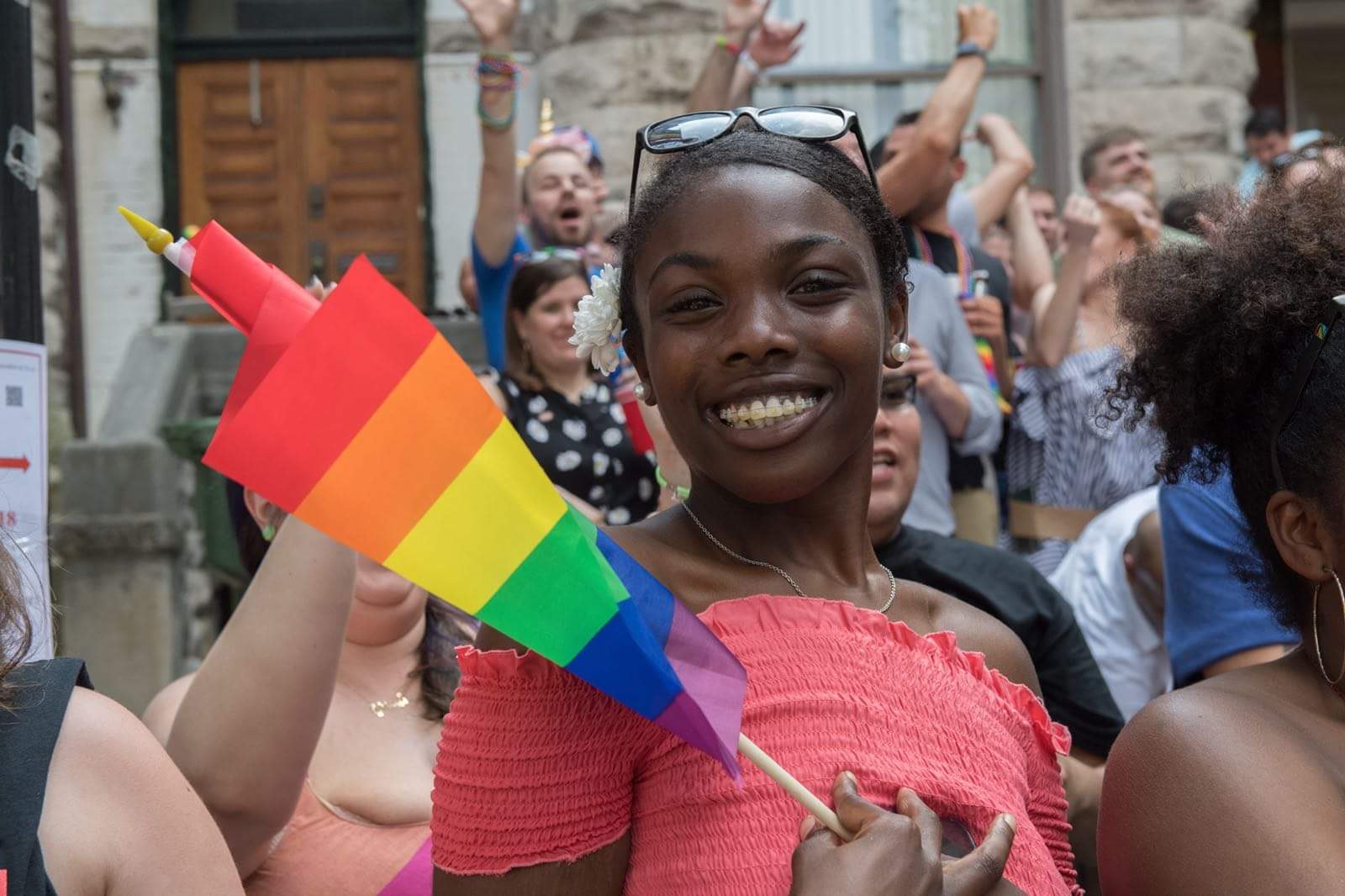 Eine Gruppe von Menschen vor einem Gebäude stehend, mit einer Frau in der Mitte, die eine Regenbogenflagge hält, wahrscheinlich an einem Christopher Street Day teilnehmend.