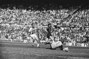 Schwarzes und weißes Foto von Fußballspielern auf einem Feld während eines Bundesliga-Spiels zwischen Ajax und Hannover 96 in der Saison 1978-79, mit Zuschauern und einer Ligatafel im Hintergrund.