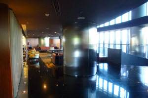 Eine geräumige Business-Lounge am Hong Kong International Airport mit großen Fenstern, hohen Decken mit elektrischen Leuchten, Sitzgruppen einschließlich Stühlen und Sofas, Tischen, einem Teppichboden und einer zentralen Säule.