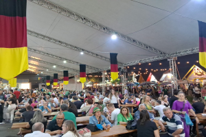 Eine große Gruppe von Menschen sitzt an langen Tischen unter einem Zelt auf dem Oktoberfest in München, Deutschland, mit einigen Stehenden und Taschen auf den Tischen, umgeben von festlichen Zelten, Lichtern und Fahnen.