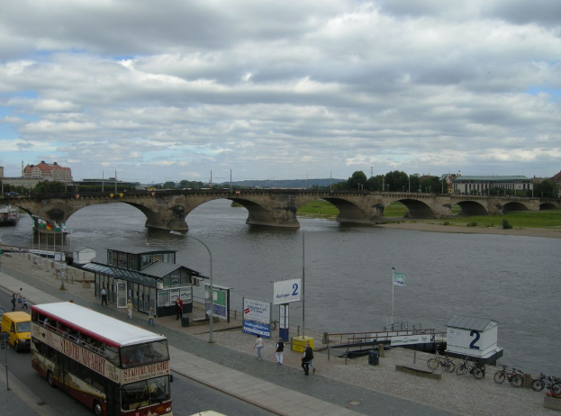 Eine belebte Straßenszene mit Fahrzeugen, Fußgängern, Fahrrädern, Gebäuden, einer Brücke und einem bewölkten Himmel, mit verschiedenen städtischen Elementen wie Plakaten, Laternen, Strommasten und Drähten.