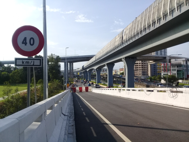 Autobahn mit Geschwindigkeitsbegrenzungsschild, Fahrzeuge, Brücke mit Pfeilern, Laternen, Bäume, Gebäude und ein bewölkter Himmel im Hintergrund.