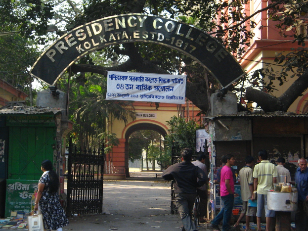Der Eingang zum Presidency College in Kolkata, Indien, mit einem Torbogen mit einem "Presidency College"-Schild, einem Tor, Menschen auf der rechten Seite, einem Gebäude, Bäumen und einem klaren blauen Himmel im Hintergrund.