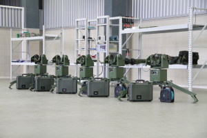 Eine Gruppe von Militärrobotern auf dem Boden umgeben von Koffern und anderen Gegenständen auf Gestellen, mit einer Wand und einem Geländer im Hintergrund.