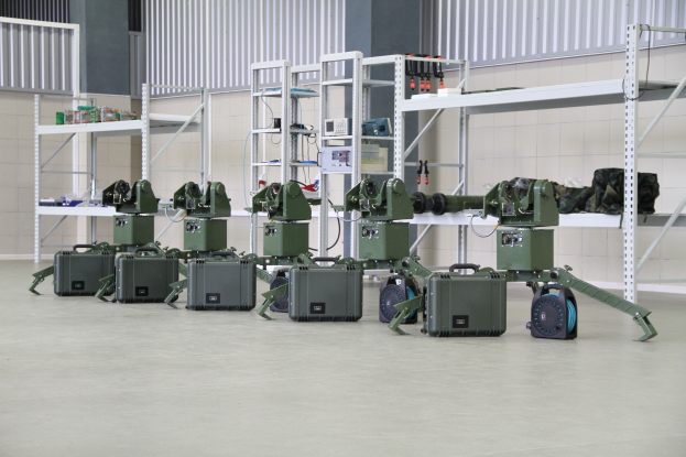 Eine Gruppe von Militärrobotern auf dem Boden umgeben von Koffern und anderen Gegenständen auf Gestellen, mit einer Wand und einem Geländer im Hintergrund.