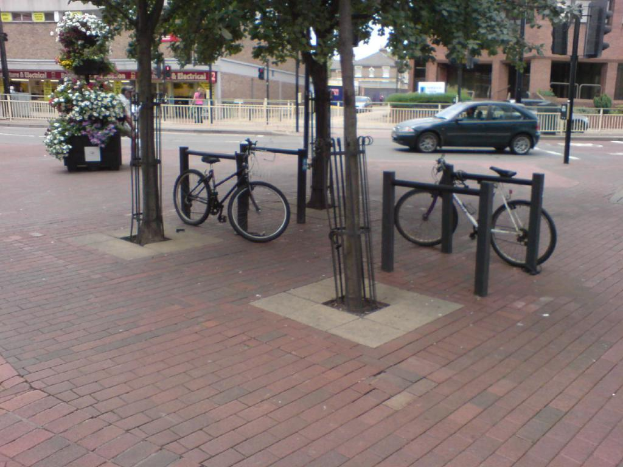 Eine Szene mit Fahrrädern, Bäumen, einem Zaun, einem Auto und Gebäuden.