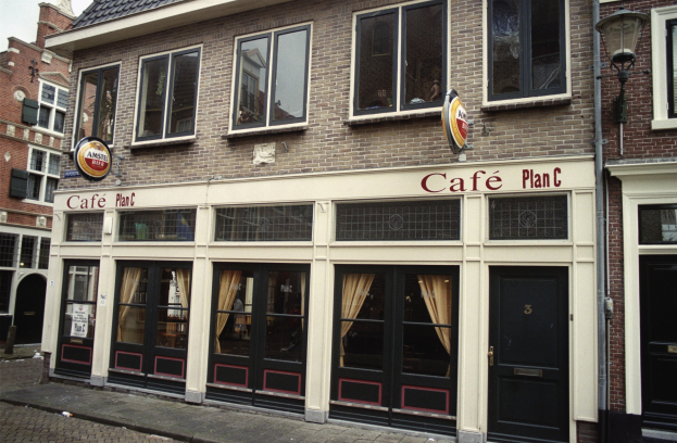 Ein Gebäude mit Fenstern und Türen, ein Schild mit der Aufschrift "Cafe Plan C in Amsterdam, Niederlande", ein Straßenpfosten, eine Baumgruppe und ein bewölkter Himmel.
