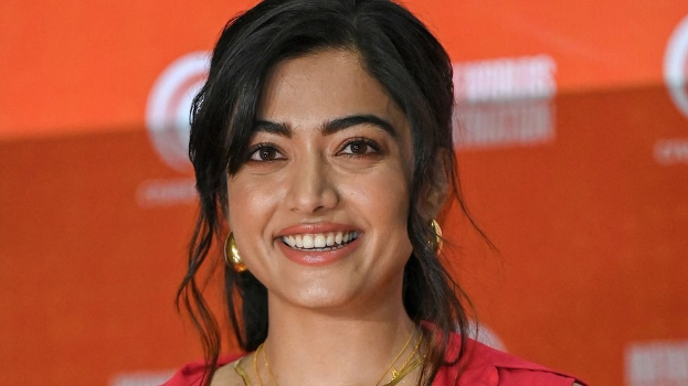 Frau in rotem Kleid mit glattem Dutt und dezentem Make-up, die in die Kamera lächelt, vor einem gelben Banner mit der Aufschrift "Sonam Kapoors beste Schönheits Looks vom Cannes Film Festival."