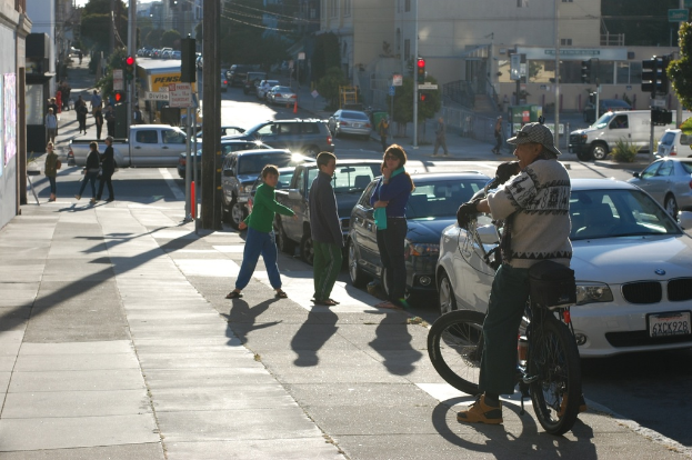 Eine belebte Straßenszene in der Stadt mit einem Bürgersteig, mehreren Fahrzeugen, Fußgängern, Ampeln, einer Person in einer Mütze und Handschuhen auf einem Fahrrad, Bäumen und Gebäuden.