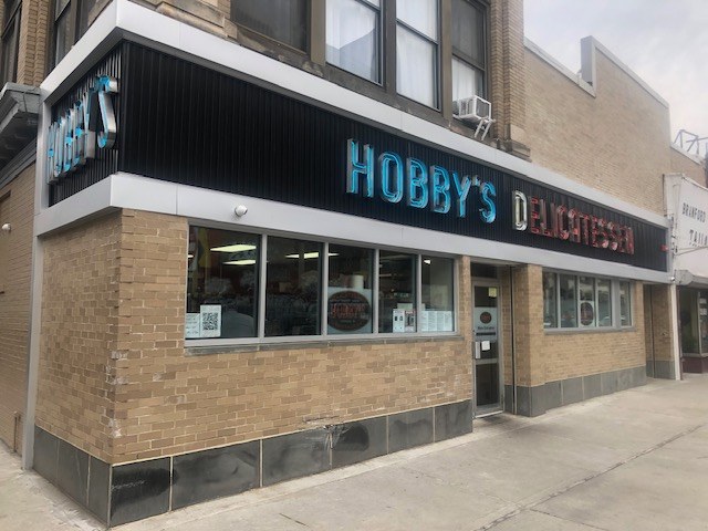 Außenansicht von Hobby's Delicatessen in Chicago, Illinois, mit Glasfenstern und -türen, einem Namensschild, einem Laternenmast davor und einem bewölkten Himmel im Hintergrund.