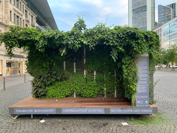Ein grünes Gebäude mit Efeu an der Seite, umgeben von Bäumen, Fahrzeugen, Polen und anderen Gebäuden, unter einem bewölkten Himmel, Teil der Staufbilter Biodiversität in Berlin, Deutschland.