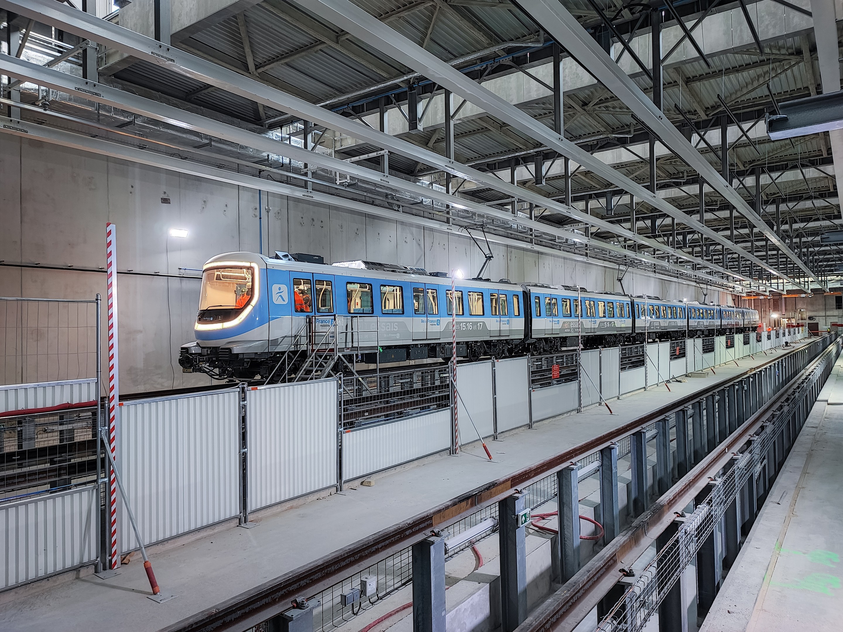 Ein blau-weißer Zug fährt durch eine große Bahnhofshalle mit einem Bahnsteig auf der rechten Seite und einer Wand im Hintergrund, unter einer Decke mit Stangen und Lampen.