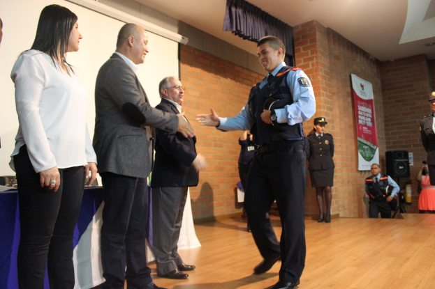 Eine Gruppe von Menschen auf einer Bühne, einer in Uniform mit einer Mütze haltend, eine Frau in einem weißten Hemd, mit einem Banner, Lautsprecher, Vorhängen, Tisch und Bildschirm im Hintergrund.