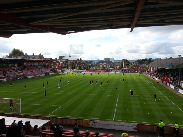 Ein Fussballspiel wird im St Mary's Stadium gespielt, mit Zuschauern auf den Bänken und Stehplätzen und einem Torpfosten links sowie Gebäuden, Bäumen und einem bewölkten Himmel im Hintergrund.