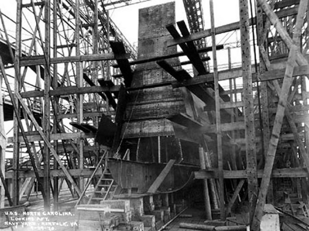 Schwarzes und weißes Foto eines Schiffs in einem Schiffbau, mit hölzernen Pfählen und Treppen im Vordergrund und Himmel im Hintergrund, mit dem Text "Marinebewaffnung" in der linken unteren Ecke.