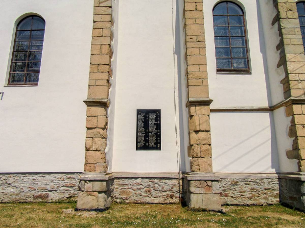 Eine weiße Kirche mit einer Steinmauer und Fenstern, umgeben von Gras und einem klaren blauen Himmel, trägt eine Gedenktafel für die Opfer des Holocaust auf ihrer Wand.
