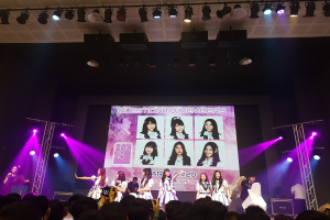 Eine Gruppe von Menschen, die auf einer hell erleuchteten Bühne spielen, mit einer Menge darunter, mit einem Sänger mit Mikrofon und einem Musiker, der ein Instrument spielt, während ein großer Bildschirm im Hintergrund Bilder der K-Pop-Gruppe BK48 zeigt.