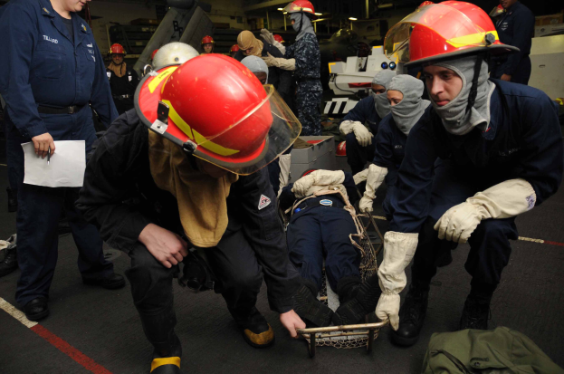 Eine Gruppe von Menschen in Helmen und Handschuhen, einer schiebend einen Wagen mit einer Person darauf, ein anderer haltend Papiere und einen Stift, mit einer Tasche auf dem Boden und verschiedenen Gegenständen im Hintergrund, auf dem Deck eines Schiffs mit Deckenbeleuchtung.
