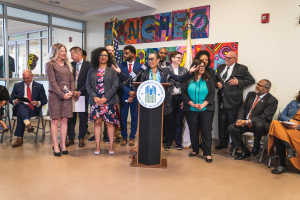 Gruppe von Menschen bei einer Stadtratveranstaltung mit einem Podium, einem Mikrofon und einer Tafel mit der Aufschrift "Sancho Stadtrat tritt dem Stadtrat bei", sitzende und stehende Teilnehmer, Flaggen, Feuerlöscher und Deckenleuchten.