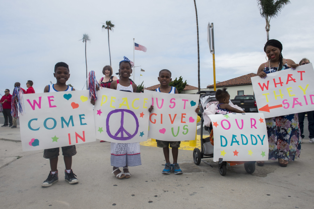 Eine Gruppe von Kindern hält Schilder in der Hand und steht vor einem Gebäude, während eine Frau einen Kinderwagen schiebt, umgeben von Bäumen, Pfählen, einer Flagge und einem bewölkten Himmel; auf einem Schild steht "Wir kommen in Frieden, gebt uns unseren Daddy."