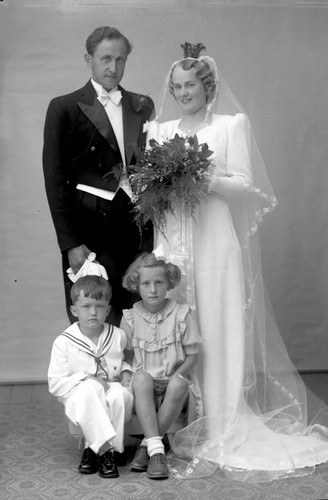 Schwarzes und weißes Foto einer Braut in einem weißen Kleid mit einem Bukett, einem grinsenden Bräutigam neben ihr und zwei Kindern, die vor ihnen auf dem Boden sitzen.