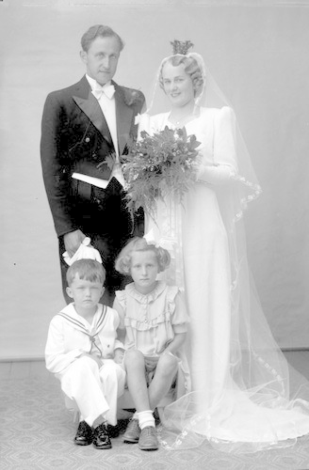 Schwarzes und weißes Foto einer Braut in einem weißen Kleid mit einem Bukett, einem grinsenden Bräutigam neben ihr und zwei Kindern, die vor ihnen auf dem Boden sitzen.
