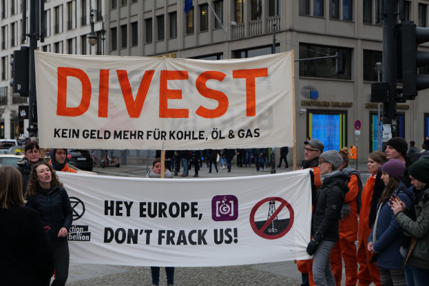 Gruppe von Menschen mit einem Banner 'Divest Hey Europe, Don't Frack Us' auf einer Straße mit Gebäuden, Straßeninfrastruktur, Fahrzeugen, Bäumen und einem klaren Himmel im Hintergrund.