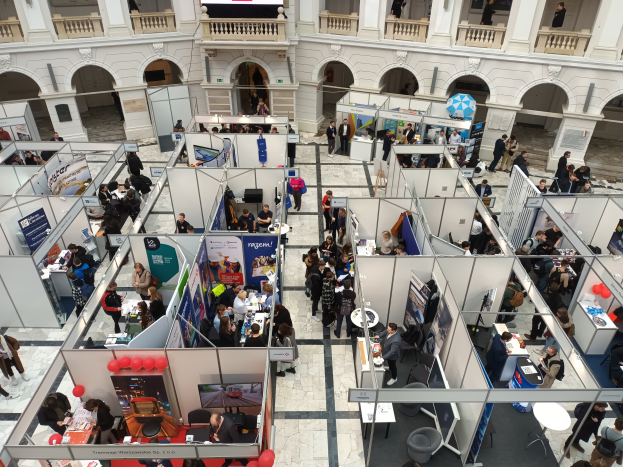 Eine große Gruppe von Menschen versammelt sich in einem Raum mit Ständen, Bannern, Tischen, Stühlen, Luftballons und einem Gebäude mit Bögen, Säulen, Geländern und einem Bildschirm im Hintergrund.