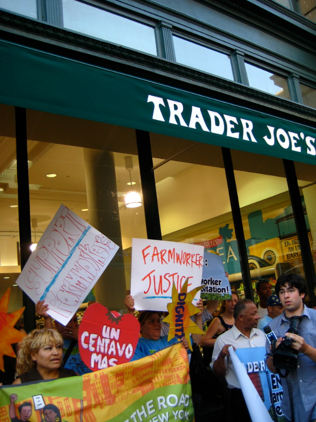 Eine Gruppe von Menschen steht in der Nähe eines Ladens und hält Schilder und Plakate, hinter ihnen ist eine Säule und Dachbeleuchtung zu sehen.