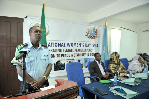 Ein Mann steht an einem Rednerpult mit einem Mikrofon und Papier, während er eine Sitzreihe mit verschiedenen Gegenständen auf dem Tisch und Flaggen, einem Banner und anderen Elementen im Hintergrund anspricht.