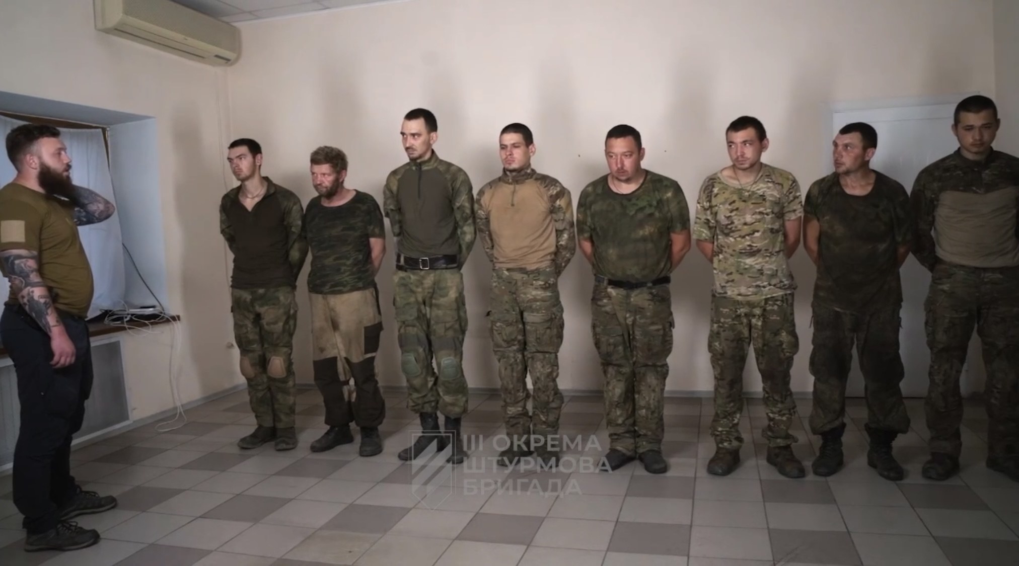 Eine Gruppe von Männern in militärischer Uniform, die zusammen in einem Raum mit einer wandmontierten Klimaanlage, einer Tür auf der rechten Seite und einem Tisch mit Gegenständen stehen; der Text unten lautet "Sergei Vorobyov und sein Team."