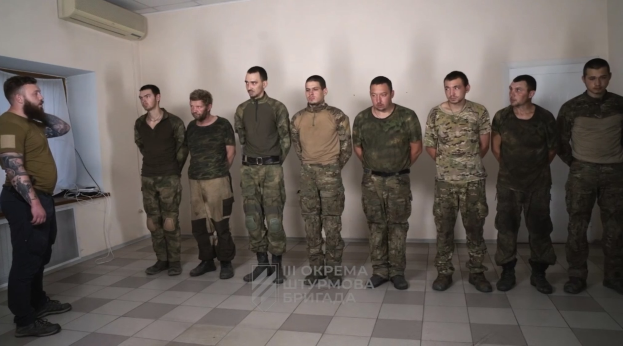 Eine Gruppe von Männern in militärischer Uniform, die zusammen in einem Raum mit einer wandmontierten Klimaanlage, einer Tür auf der rechten Seite und einem Tisch mit Gegenständen stehen; der Text unten lautet "Sergei Vorobyov und sein Team."