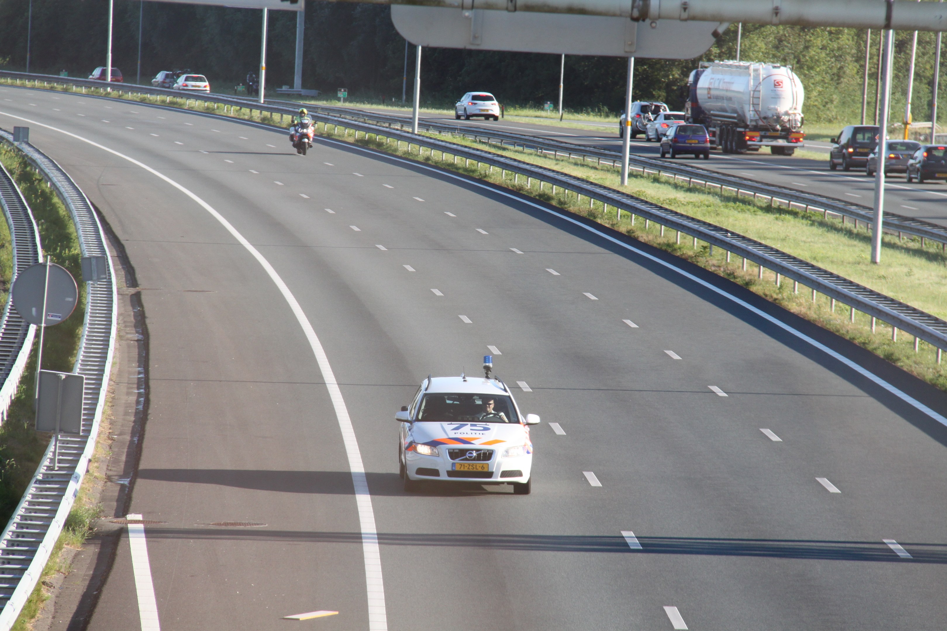 Ein Auto auf einer Autobahn mit einem stationären Polizeiwagen am Straßenrand, andere Fahrzeuge und Bäume, Pfosten und Geländer im Hintergrund.