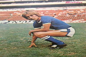 Ein Mann kniet auf einem Fußballfeld umgeben von Menschen mit Schildern auf Stühlen und Tafeln, trägt sportliche Schuhe.