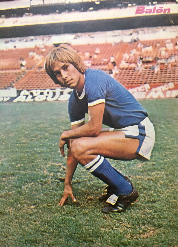 Ein Mann kniet auf einem Fußballfeld umgeben von Menschen mit Schildern auf Stühlen und Tafeln, trägt sportliche Schuhe.