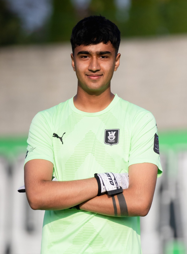Ein junger Mann in einem grünen Hemd auf einem Fußballfeld mit verschränkten Armen und einem Lächeln, mit einem unscharfen Hintergrund.