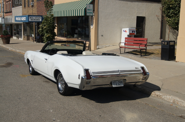 Ein weißes Auto mit der Nummer 771-299 steht auf der Straße, rechts daneben eine Bank, im Hintergrund ein Gebäude und ein Geschäft namens 'Immobilienversicherung'.