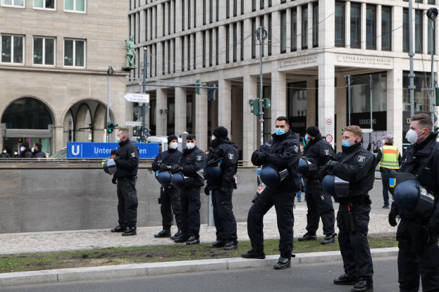 Eine Gruppe von Polizisten in schwarzen Uniformen und Masken steht vor einem Glasfenstergebäude mit Säulen in Berlin, Deutschland, einige halten Helme, mit Laternenmasten, Verkehrszeichen, Texttafeln, einer Statue, anderen Menschen und grasbewachsenem Boden im Hintergrund.