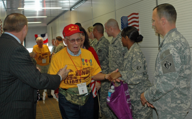 Eine Gruppe von Menschen und Soldaten, die drinnen mit einer Flagge an der rechten Wand und Lichtern an der Decke stehen.